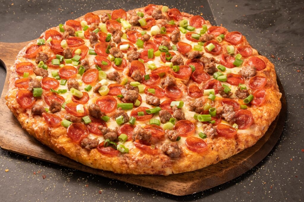 Photo of a combination pizza topped with pepperoni slices, sausage, and green onions. The crust is golden brown and crispy. The pizza is resting on a wooden paddle as though it just came out of the oven. At Me-N-Ed's Coney Island Grill in Fresno, CA, you can choose from our delicious signature pizzas or create your own!